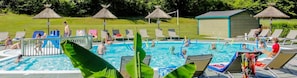 Una piscina con sillones de descanso y un tobogán, rodeada de césped y un pequeño edificio.