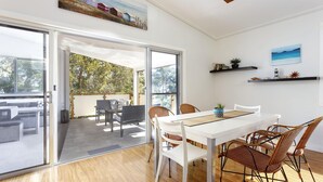 Un moderno comedor con una mesa y sillas blancas, una puerta de vidrio que da a un patio exterior y vistas al exterior.