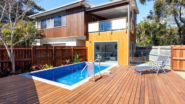 Una casa de dos plantas con terraza de madera, piscina y patio amoblado.