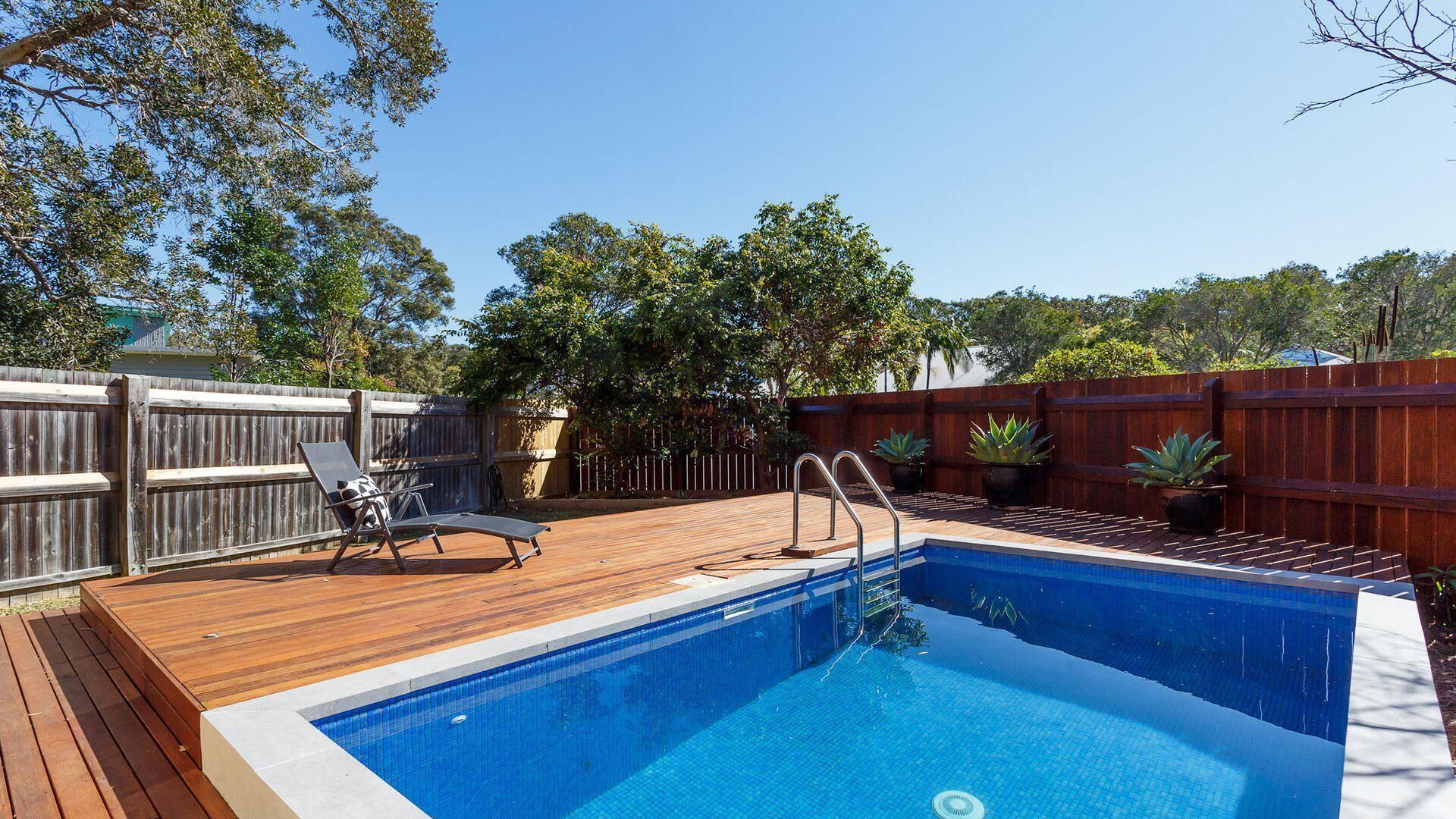 A swimming pool with a wooden deck, a lounge chair, and a wooden fence.