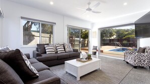 Una sala moderna con un sofá gris, una mesa de centro blanca y sillones estampados. Amplias ventanas y una puerta corrediza de vidrio dan acceso a una zona de piscina.