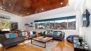 Un salón espacioso con techo de madera, ventanas amplias que ofrecen vistas a árboles y casas, dos sofás grises con cojines estampados y una mesa de centro.