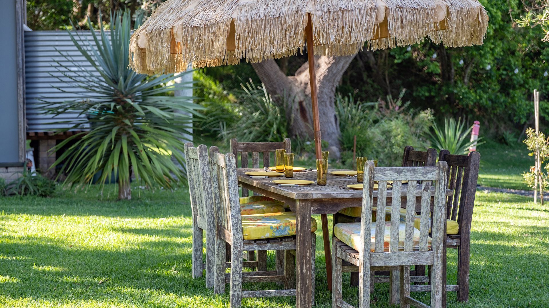 A rustic wooden table with a thatched umbrella, surrounded by weathered wooden chairs with yellow cushions, set in a lush garden.