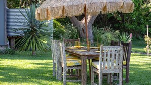 A rustic wooden table with a thatched umbrella, surrounded by weathered wooden chairs with yellow cushions, set in a lush garden. - Pippis - a Classic Beach House (Seal Rocks)