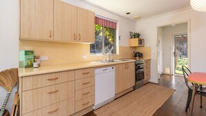 A modern kitchen with wooden cabinets, a dishwasher, a stove, and a window with a striped valance. - Pippis - a Classic Beach House (Seal Rocks)