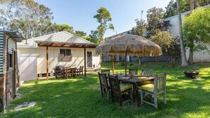 Ein Garten mit einem Holztisch und Stühlen, einem Reetdachschirm und einem kleinen Haus mit Garage.