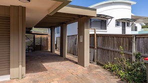 A covered outdoor area with a brick floor, wooden fence, and a building with a white exterior and windows.