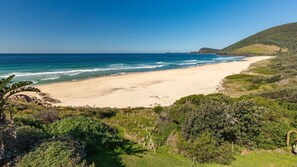 A coastal landscape with a sandy beach, greenery, and a clear sky.