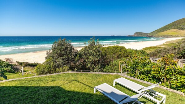 A beach view with a grassy area, two white lounge chairs, and a wooden fence.