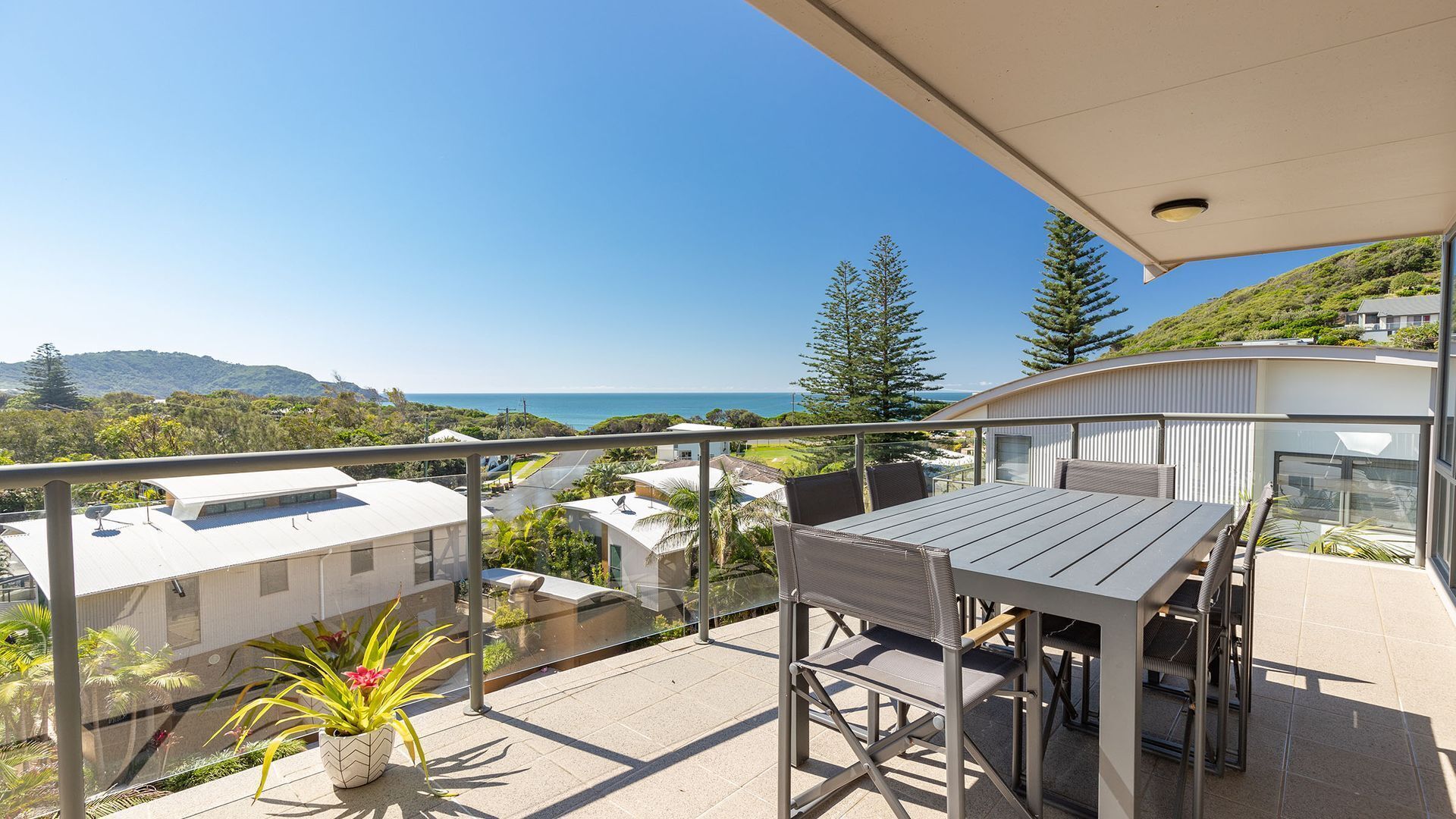 A balcony with a table and chairs, overlooking a coastal landscape with buildings and greenery.