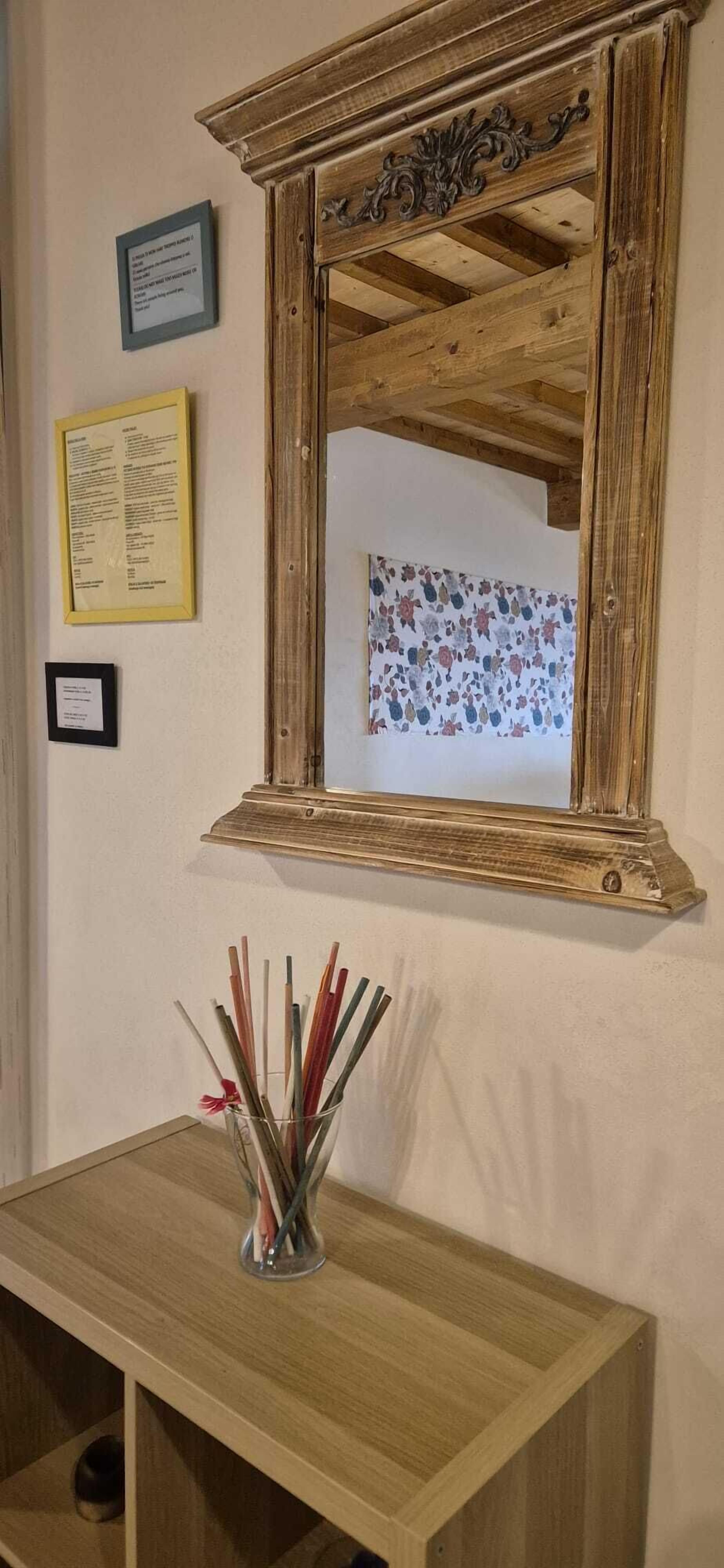 A mirror with a wooden frame, a small table with a vase of reeds, and framed informational plaques on the wall.