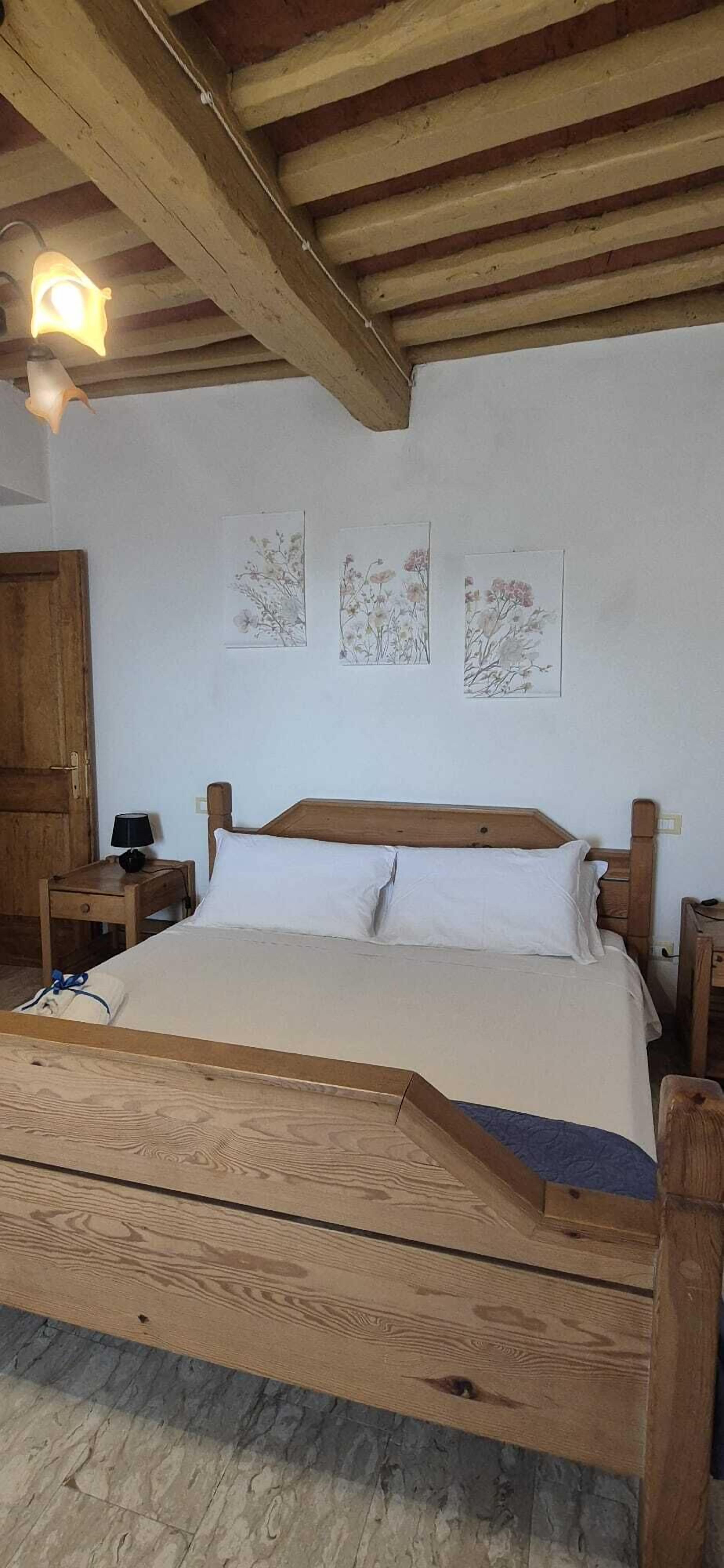 Een bed met houten frame, een witte matras en kussens, een nachtkastje met een lamp, en drie ingekaderde botanische prints aan de muur.