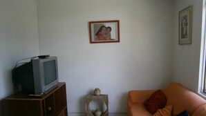 A living room with a television on a wooden cabinet, a small table with a vase, and an orange couch with pillows. - Servicio de Hotel (Bogotá)