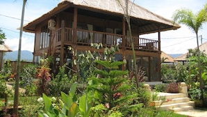 A two-story wooden house with a thatched roof, surrounded by lush tropical vegetation.