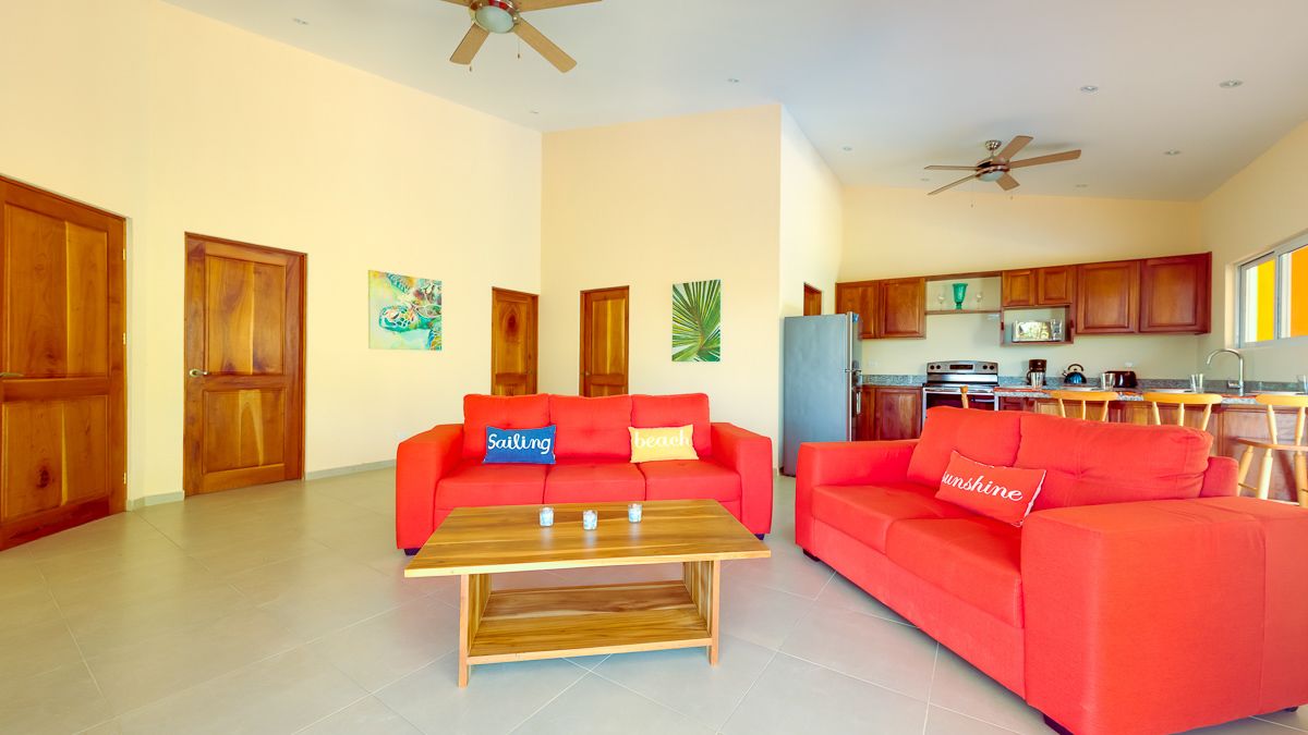 A spacious living room with a red sofa, a wooden coffee table, and a kitchen area in the background.