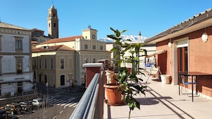 Ein Balkon mit Blumentöpfen, einem Tisch und Stühlen mit Blick auf eine Straße, auf der Autos geparkt sind und Gebäude stehen, darunter ein im Hintergrund sichtbarer Glockenturm.