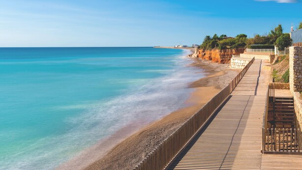 Ein Küstenweg entlang eines Strandes mit klarem, blauem Wasser und einer Steinmauer.