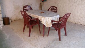 Une table ronde recouverte d’une nappe, entourée de quatre chaises en bois, dans une pièce dont le mur présente une texture particulière et où se trouve une poubelle.