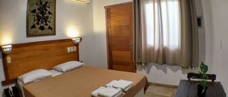 A hotel room with a wooden bed, a nightstand, a bedside lamp, a wall-mounted air conditioner, a door, and a window with curtains.
