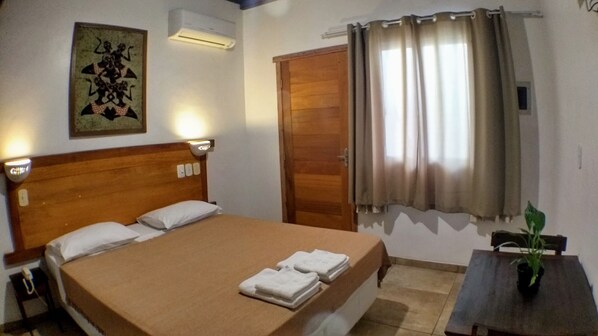 A hotel room with a wooden bed, a nightstand, a bedside lamp, a wall-mounted air conditioner, a door, and a window with curtains.