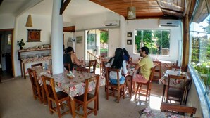 A dining area with wooden tables and chairs, a kitchen in the background, and a view of greenery outside. - Pousada Pé na Estrada (Porto Seguro)