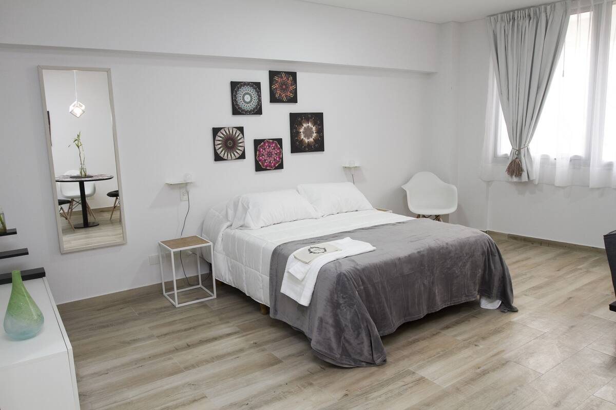Ein modernes Schlafzimmer mit Bett, Nachttischen, einem Spiegel, Wandbildern und einem Fenster mit Vorhängen.