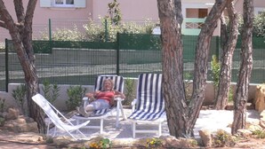 Eine Person entspannt auf einer gestreiften Liege in einem sonnigen Außenbereich mit Bäumen und einem Zaun im Hintergrund.