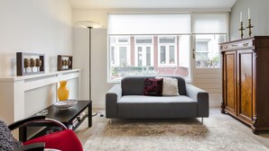 Een woonkamer met een grijze bank, een houten kast en een raam met uitzicht op een bakstenen gebouw.