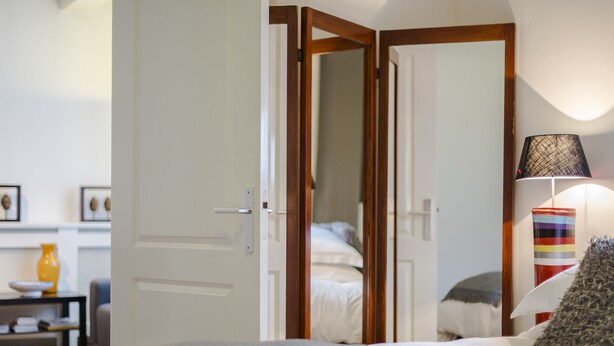 Een slaapkamer met een bed, een bedlampje, een spiegel en een deur naar een andere kamer.