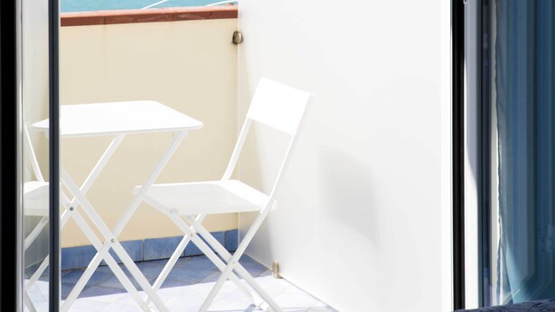 A balcony with a view of the sea, white folding chairs, and a bed.