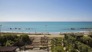 A coastal view with a paved walkway leading to the beach, surrounded by greenery and a clear sky.