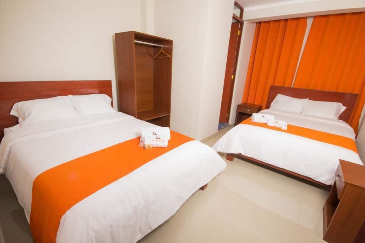 Habitación de hotel con dos camas, cada una con ropa de cama blanca y detalles naranja, un armario de madera y una cortina naranja.