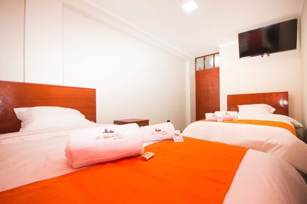 Habitación de hotel con dos camas, cada una con cabecero de madera, ropa de cama blanca y colchas anaranjadas. Hay un televisor de pantalla plana montado en la pared sobre las camas.