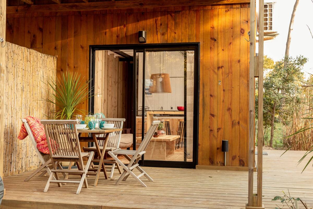 Una terraza de madera con una mesa, sillas, una lámpara colgante y un ventanal corredera que da a un espacio interior.