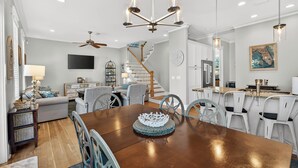 Une salle à manger spacieuse avec une table en bois, des chaises blanches et un lustre. Un escalier mène à l’étage supérieur, et on aperçoit une cuisine au fond.
