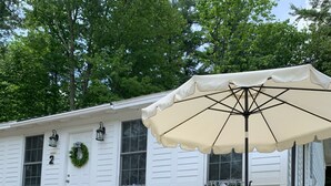Un espace extérieur accueillant avec des sièges confortables, une maison blanche en arrière-plan, un jardinier en bois orné de fleurs roses et une enseigne indiquant « HUILE MOTEUR SULONY ».