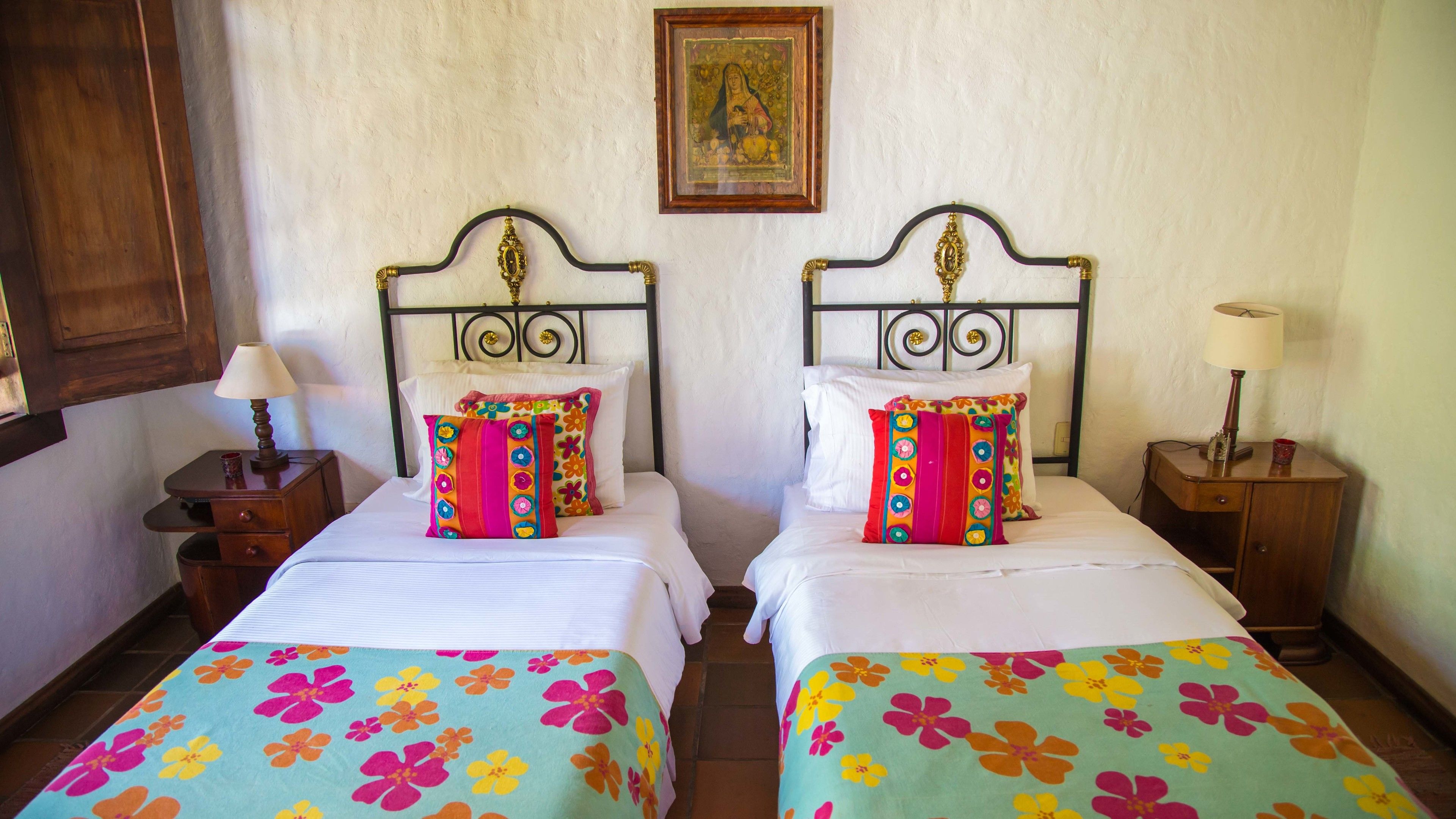 Una habitación de hotel con dos camas, cada una con cubrecamas y almohadas de colores y motivos florales.  A cada lado de las camas hay una mesita de noche de madera con una lámpara, y un cuadro enmarcado en la pared sobre las camas.