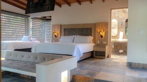 Ein Schlafzimmer mit zwei Betten, einem Holz-Kopfteil, einem Nachttisch, einer Bank und einem durchgehenden Türspalt einsehbarer Badezimmer.