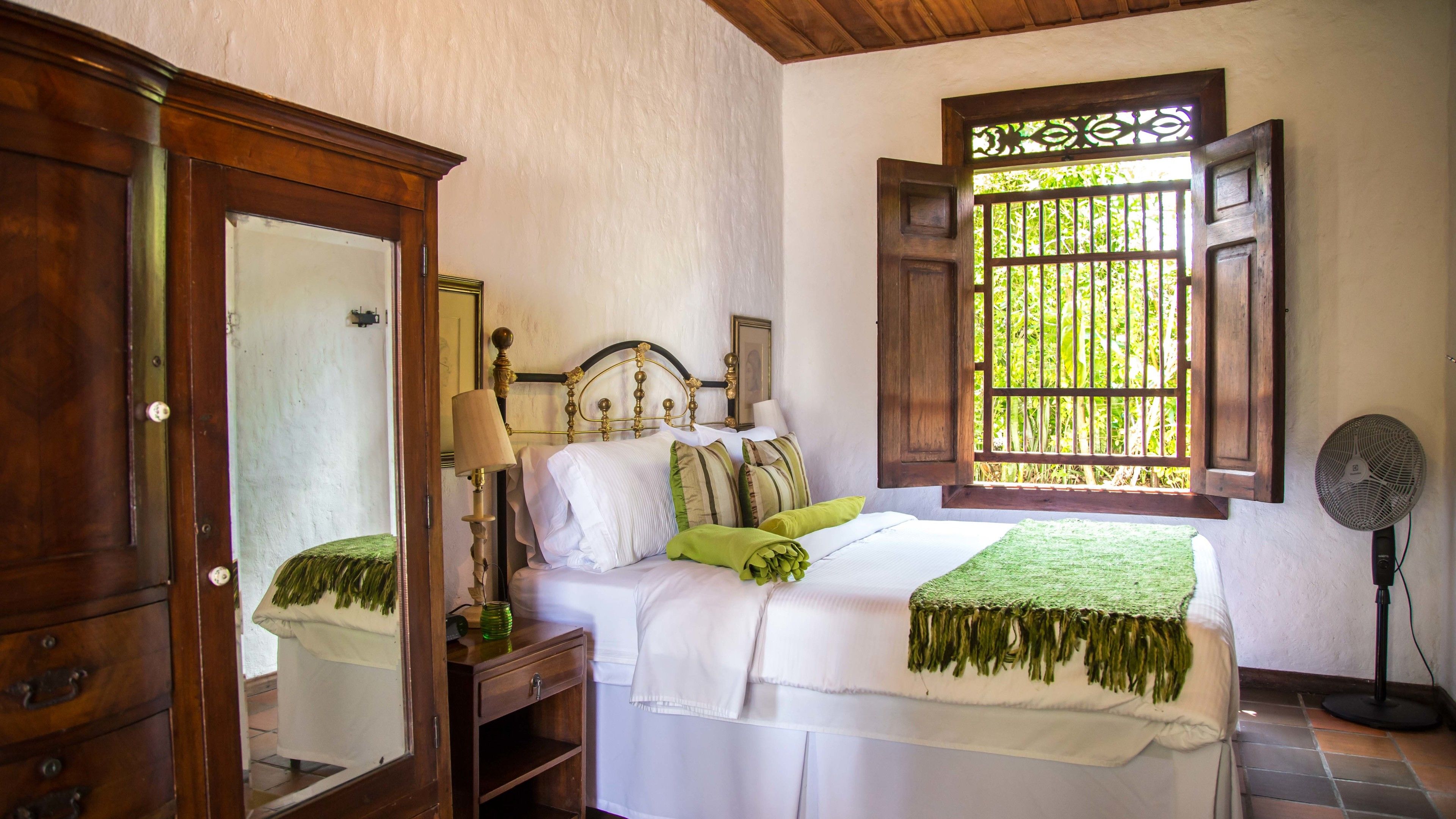 Un dormitorio con cama, armario de madera, mesita de noche con lámpara, ventana con vista a vegetación y ventilador de pie.