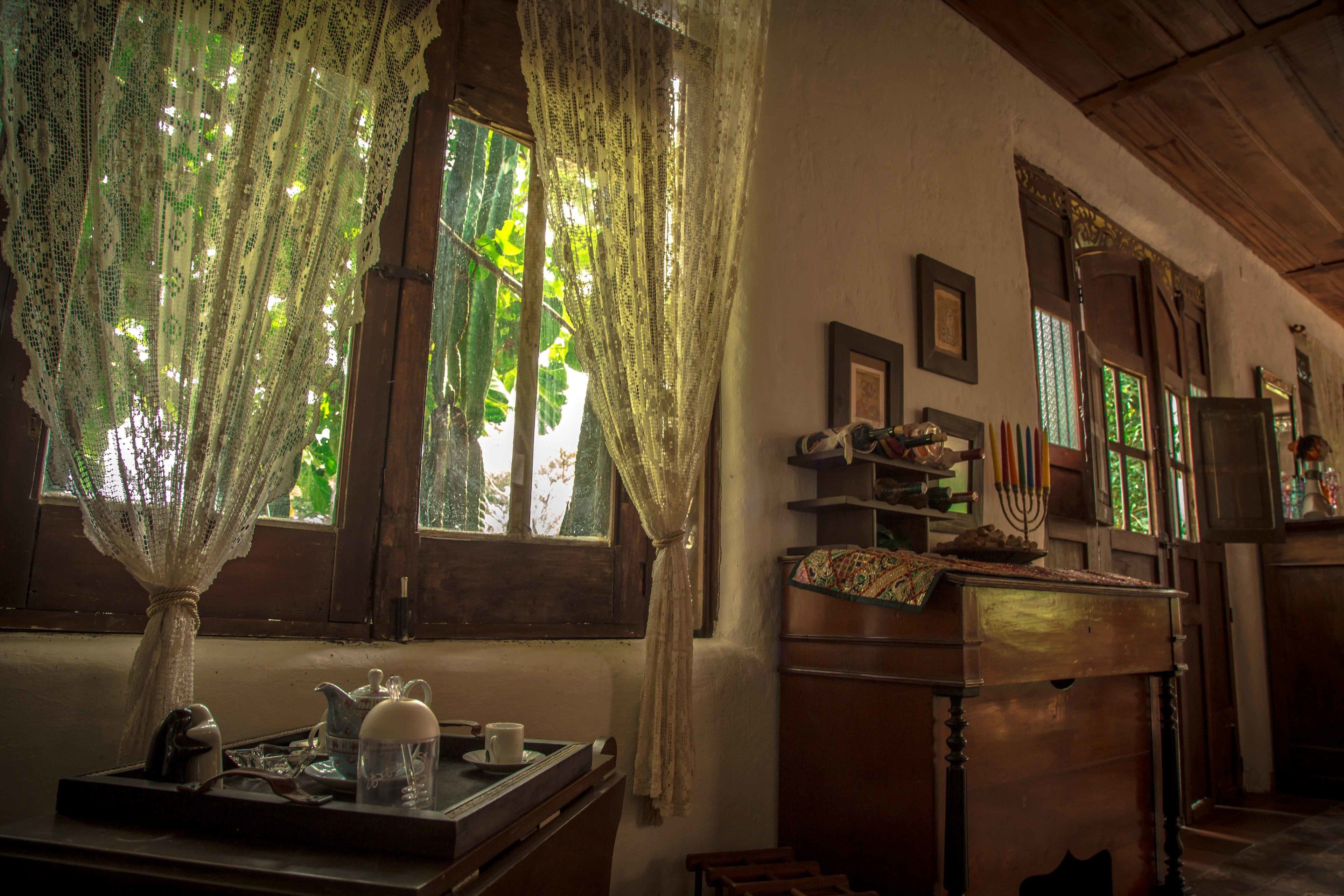 A cozy room with a wooden table, a window with lace curtains, and a shelf with various items.