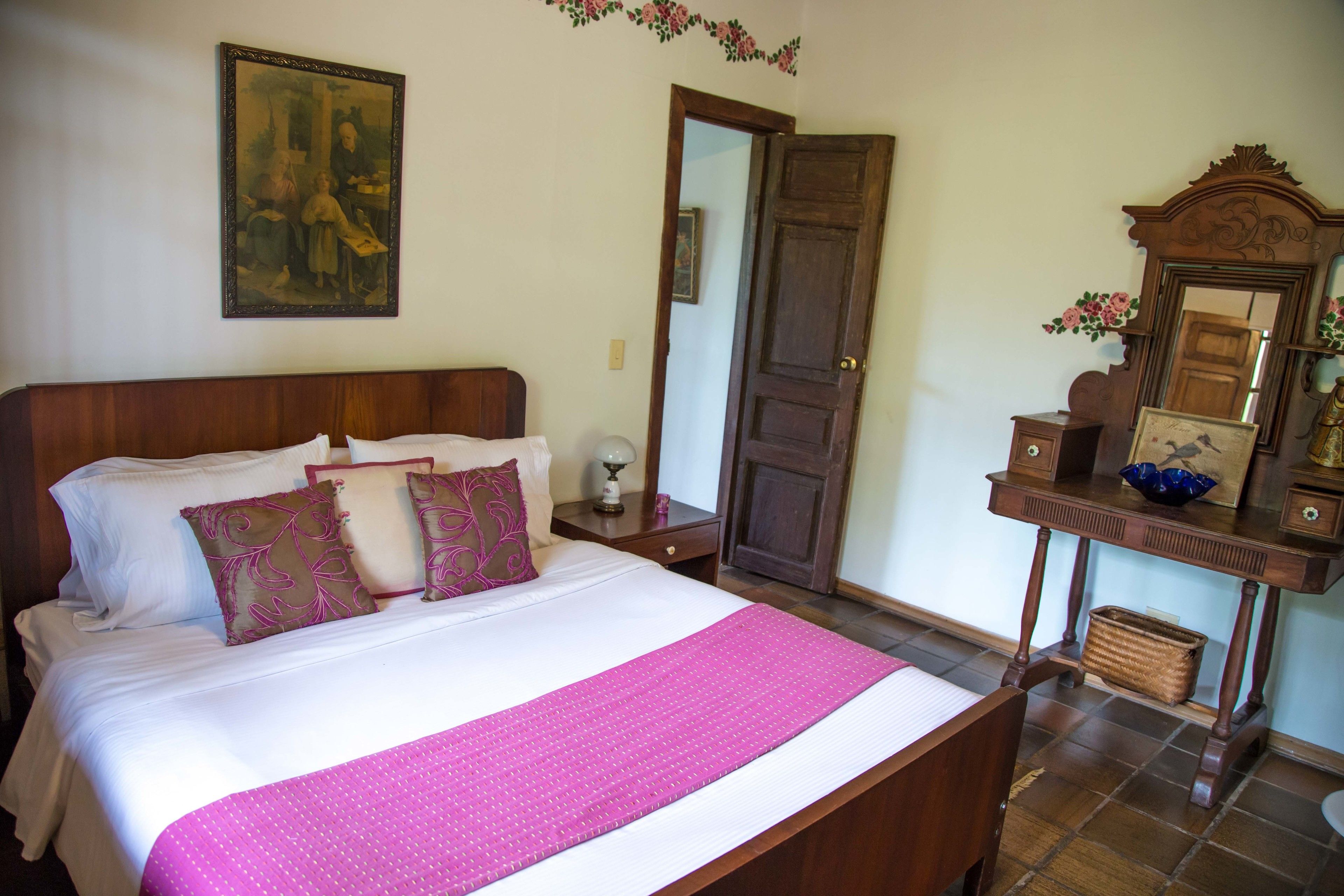 A neatly made bed with a pink blanket, a wooden nightstand with a lamp, a dresser with a mirror, and a painting on the wall.