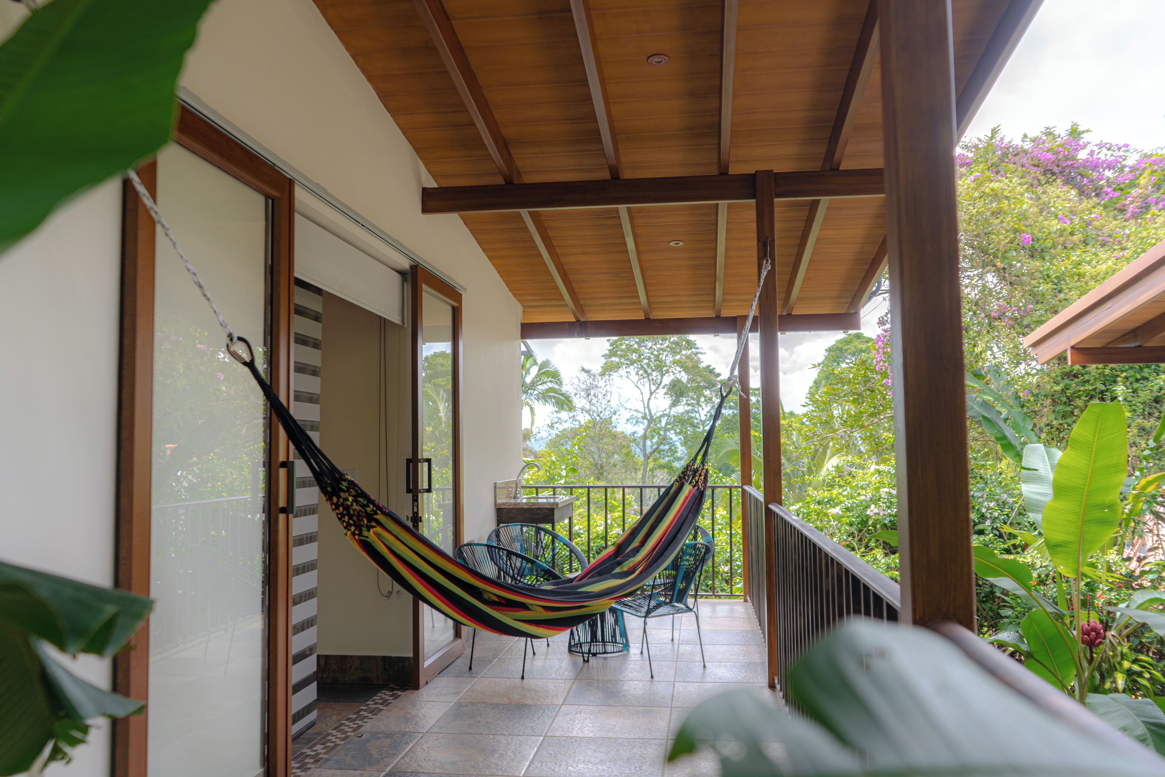 A covered balcony with a hammock, chairs, and a view of the garden.
