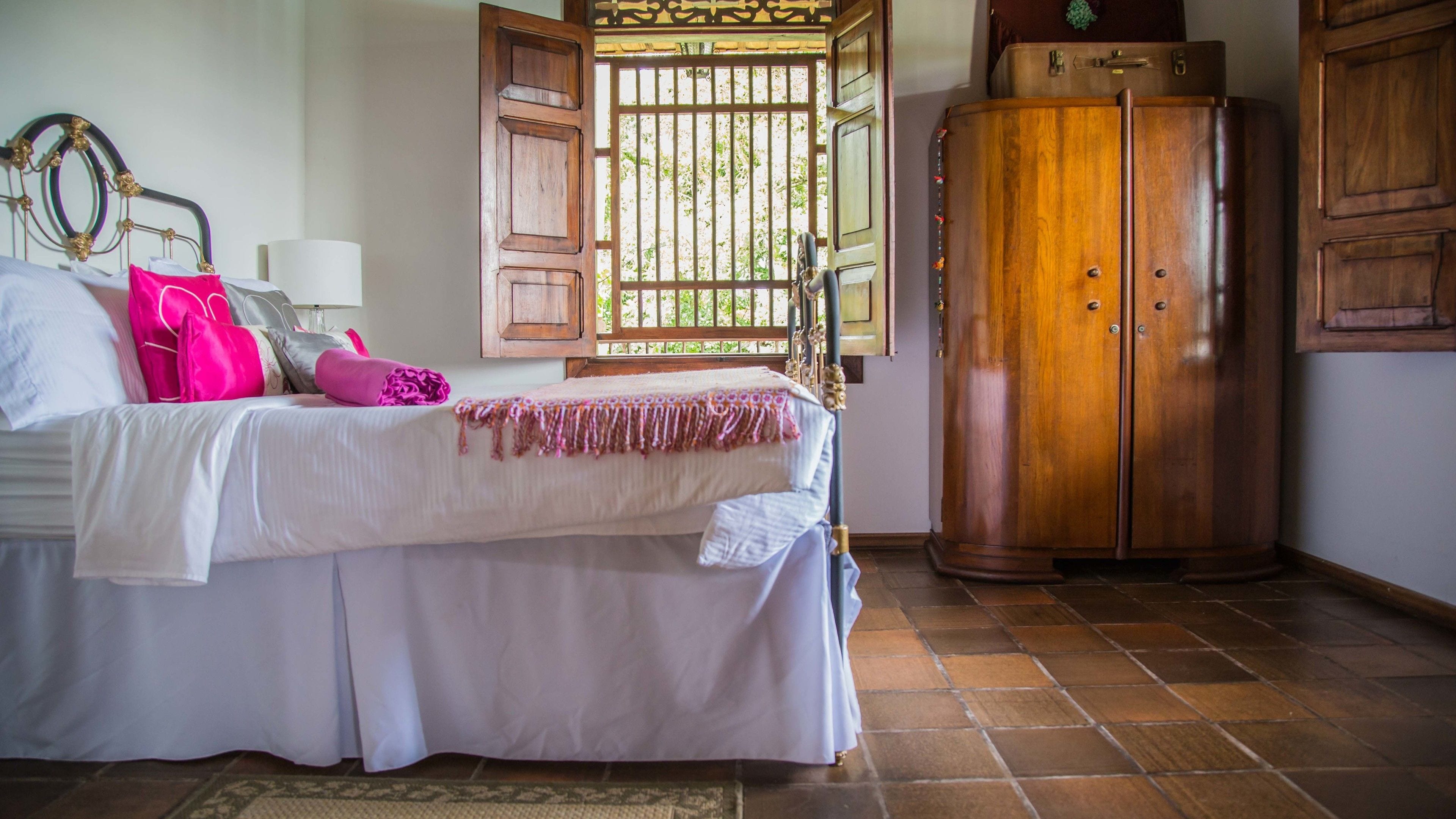 A bed with a white bedspread, pink pillows, and a wooden wardrobe in a room with tiled flooring.
