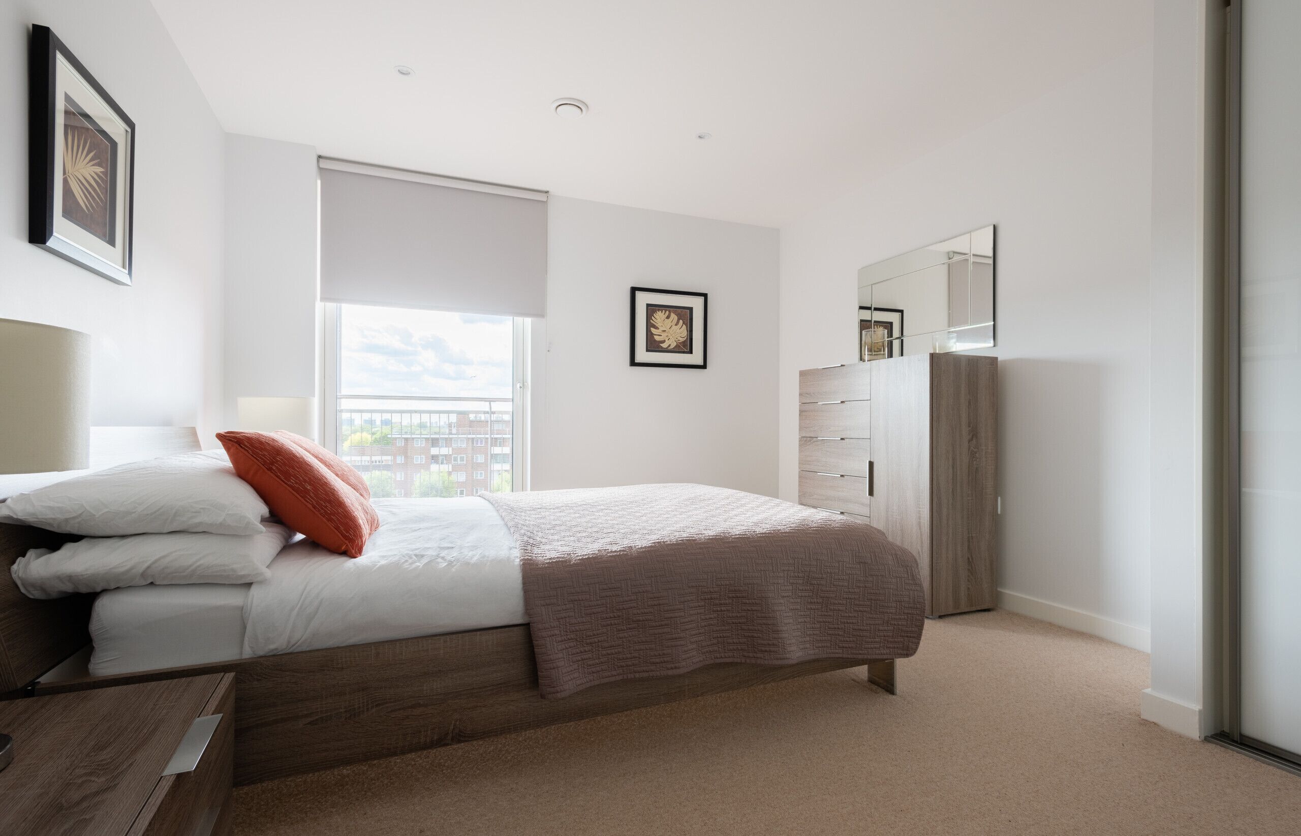 A modern bedroom with a large bed, a dresser, a mirror, and a window with a view of buildings.