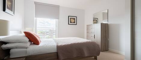 A modern bedroom with a large bed, a dresser, a mirror, and a window with a view of buildings.