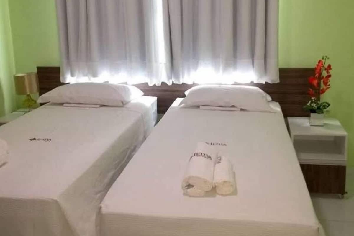 A hotel room with two beds, a nightstand, a small table with a plant, and a window with curtains.