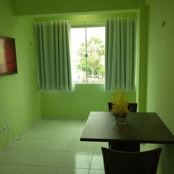 A room with a green wall, a window with curtains, a tiled floor, a table with a vase of flowers, and a chair. - Letiva Hotel (Sobral)
