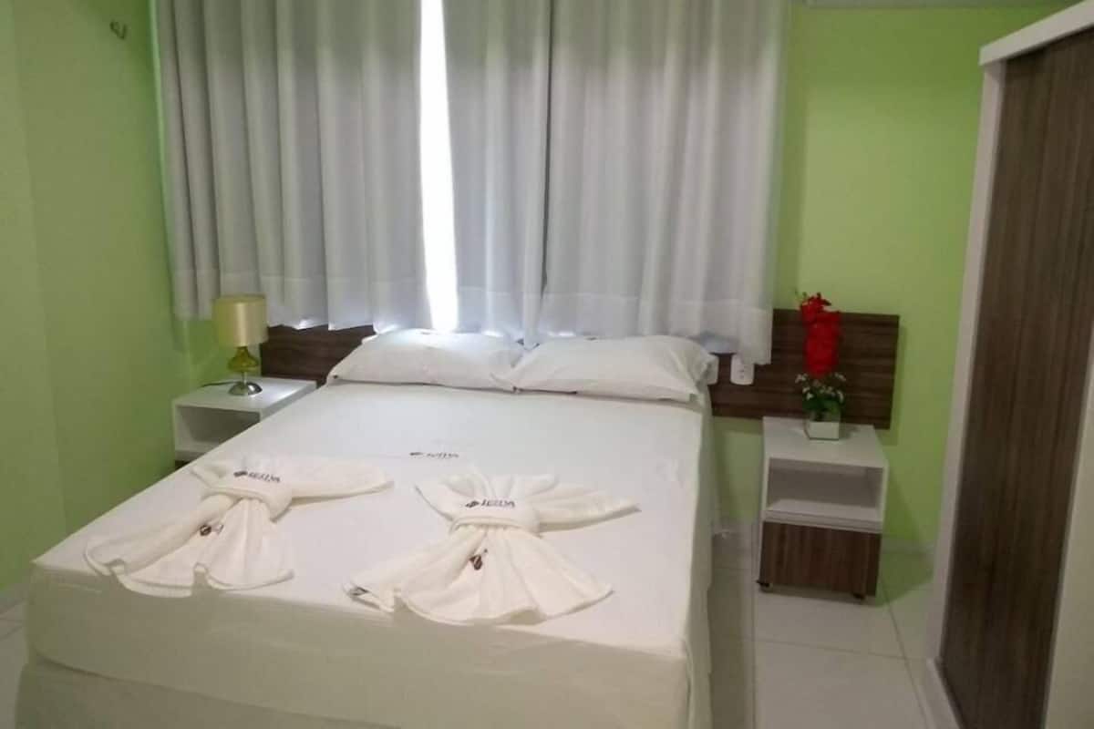 A hotel room with a bed, a nightstand, a lamp, a window with curtains, and a wall-mounted air conditioner.