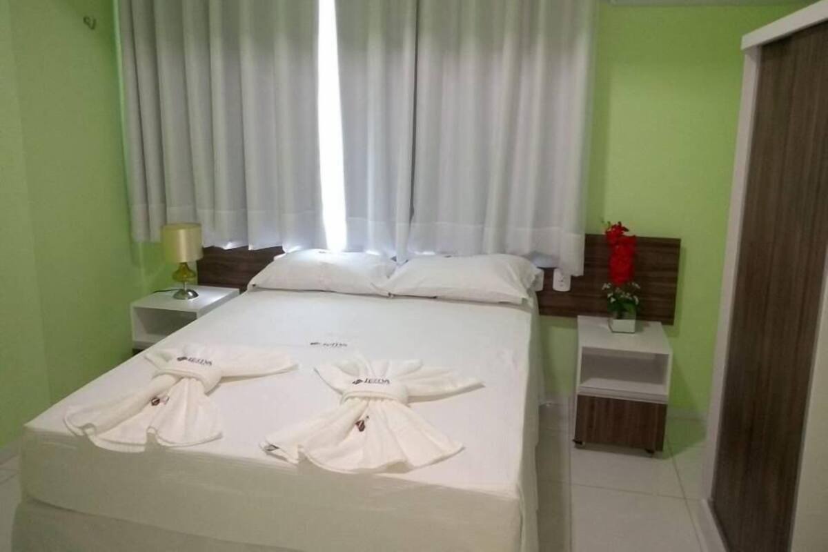 A hotel room with a bed, a nightstand, a lamp, a window with curtains, and a wall-mounted air conditioner.