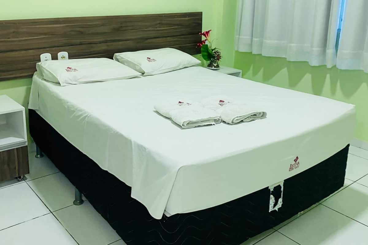 A neatly made bed with white linens and two towels, a wooden headboard, a bedside table with a lamp, and a window with white curtains.